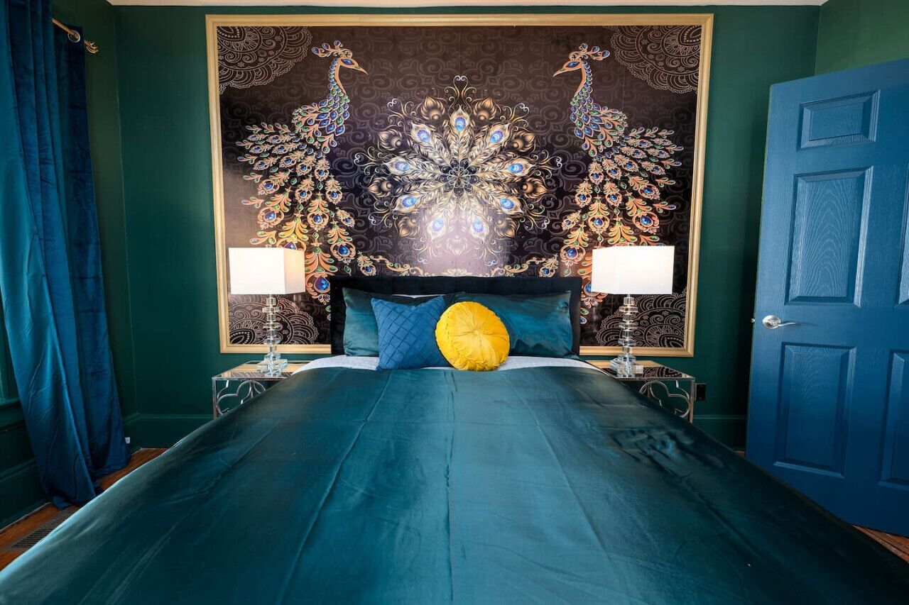 A bed with a teal bedspread, two lamps on either side, and a peacock-themed tapestry on the wall.