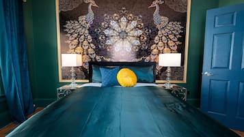 A bed with a teal bedspread, two lamps on either side, and a peacock-themed tapestry on the wall.