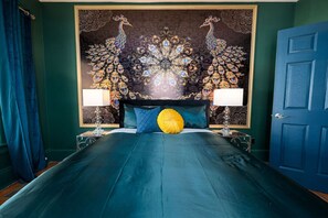 A bed with a teal bedspread, two lamps on either side, and a peacock-themed tapestry on the wall.