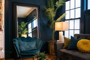 A room with a dark blue wall, a large mirror, a blue armchair with patterned cushions, a brown sofa with yellow and blue cushions, and a tall potted plant.