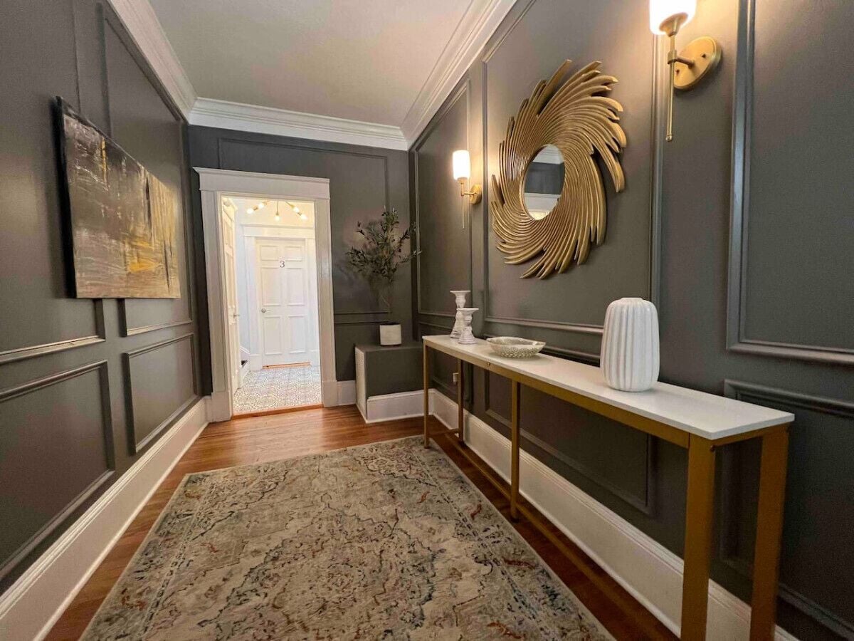 Un couloir orné d’un tapis à motifs, une console sur laquelle est posé un vase, un miroir et une porte donnant sur une autre pièce.