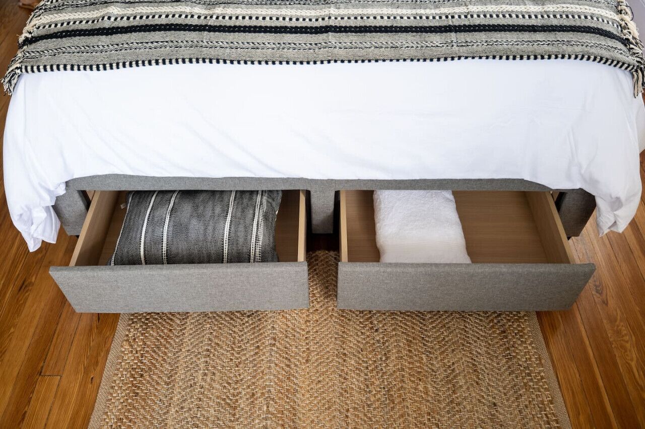 A bed with a white sheet and a striped pillow, a grey blanket, and a woven rug on a wooden floor.