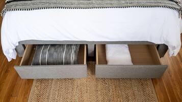 A bed with a white sheet and a striped pillow, a grey blanket, and a woven rug on a wooden floor.