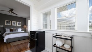 Een slaapkamer met een bed, een zwarte koelkast, een raam met zonwering en een plank met servies.
