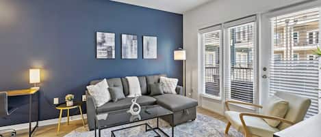 A modern living room with a grey sofa, a round coffee table, a side table with a lamp, and a large window with blinds.