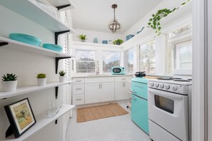 Een keuken met witte kastjes, een blauwgroene kookplaat en een magnetron. Op de planken staan planten in potten en er hangt een plant.