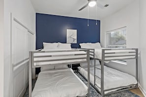 Une chambre avec un lit superposé, un ventilateur de plafond, une fenêtre avec des stores et un tableau au mur.