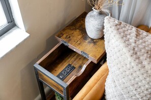 Una mesita de noche de madera con un cajón, un florero con plantas secas y una manta sobre la cama.