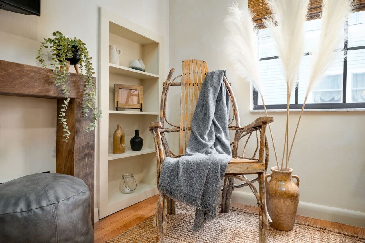 Una silla de madera rústica con un plaid gris, un tapete tejido y un jarrón con plantas secas.