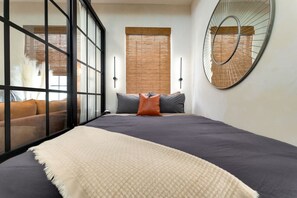 A bedroom with a large bed, a sofa, a mirror, and a window with blinds.