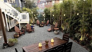 Una terraza de madera con una mesa y sillas, rodeada de plantas en macetas y un pozo para fuego.