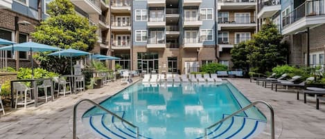 Una piscina con sillas de descanso y sombrillas, rodeada de edificios residenciales de varios pisos.