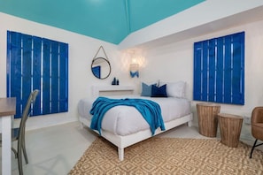 Una camera da letto con un letto bianco, dettagli blu, uno specchio rotondo e un punto luce fissato al muro.