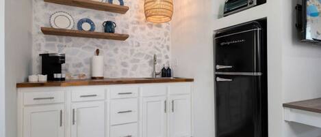 Una cocina con armarios blancos, un refrigerador negro, estantes de madera con vajilla y una lámpara colgante.