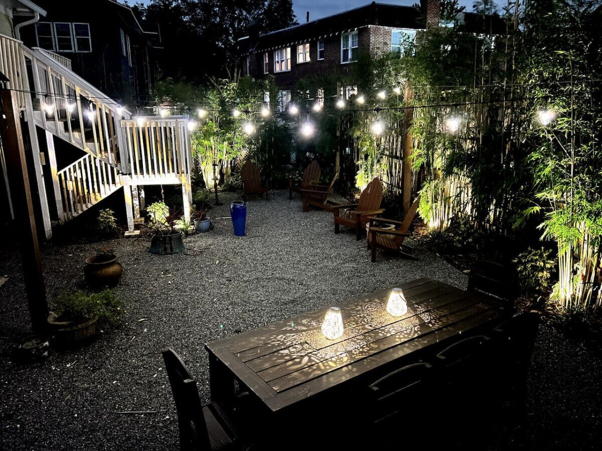 Una zona patio con tavolo e sedie di legno, circondata da piante in vaso e luci decorative.