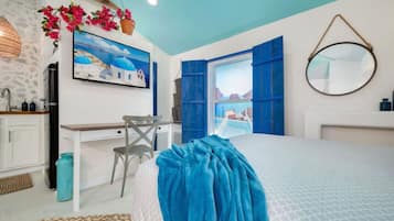 A bedroom with a bed, a desk, a chair, a window with blue shutters, a painting of a coastal scene, and a round mirror on the wall.