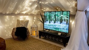 Ein gemütliches Zelt mit einem Fernseher, der eine Waldszene zeigt, ein Korbsessel, eine Laterne und ein weißer Vorhang.