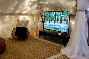 Ein gemütliches Zelt mit einem Fernseher, der eine Waldszene zeigt, ein Korbsessel, eine Laterne und ein weißer Vorhang.