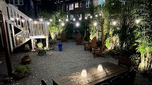 Een terras buiten met een houten tafel en stoelen, omringd door plantenpotten en lichtslingers.