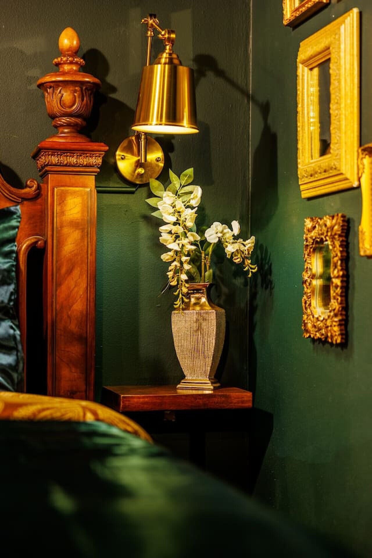 A room with a green wall, a wooden sideboard, a brass lamp, a vase with flowers, and framed pictures.