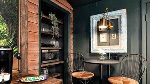 Ein gemütlicher Essbereich mit einem Holztisch und zwei Metallstühlen, einem kleinen runden Tisch mit Lampe sowie einem wandmontierten Regal mit Gläsern und Pflanzen.
