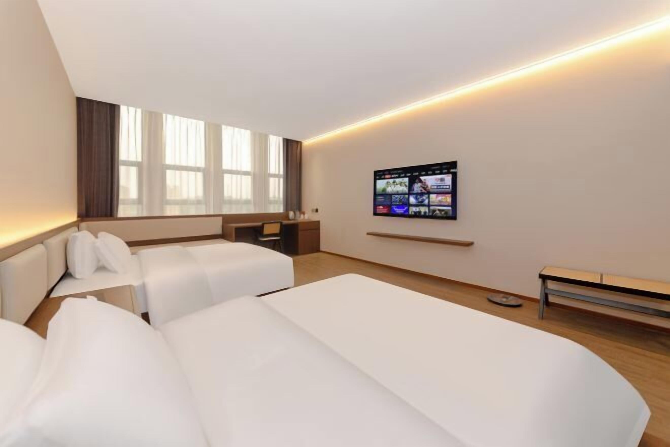 A hotel room with two beds, a desk, a TV mounted on the wall, and a large window.