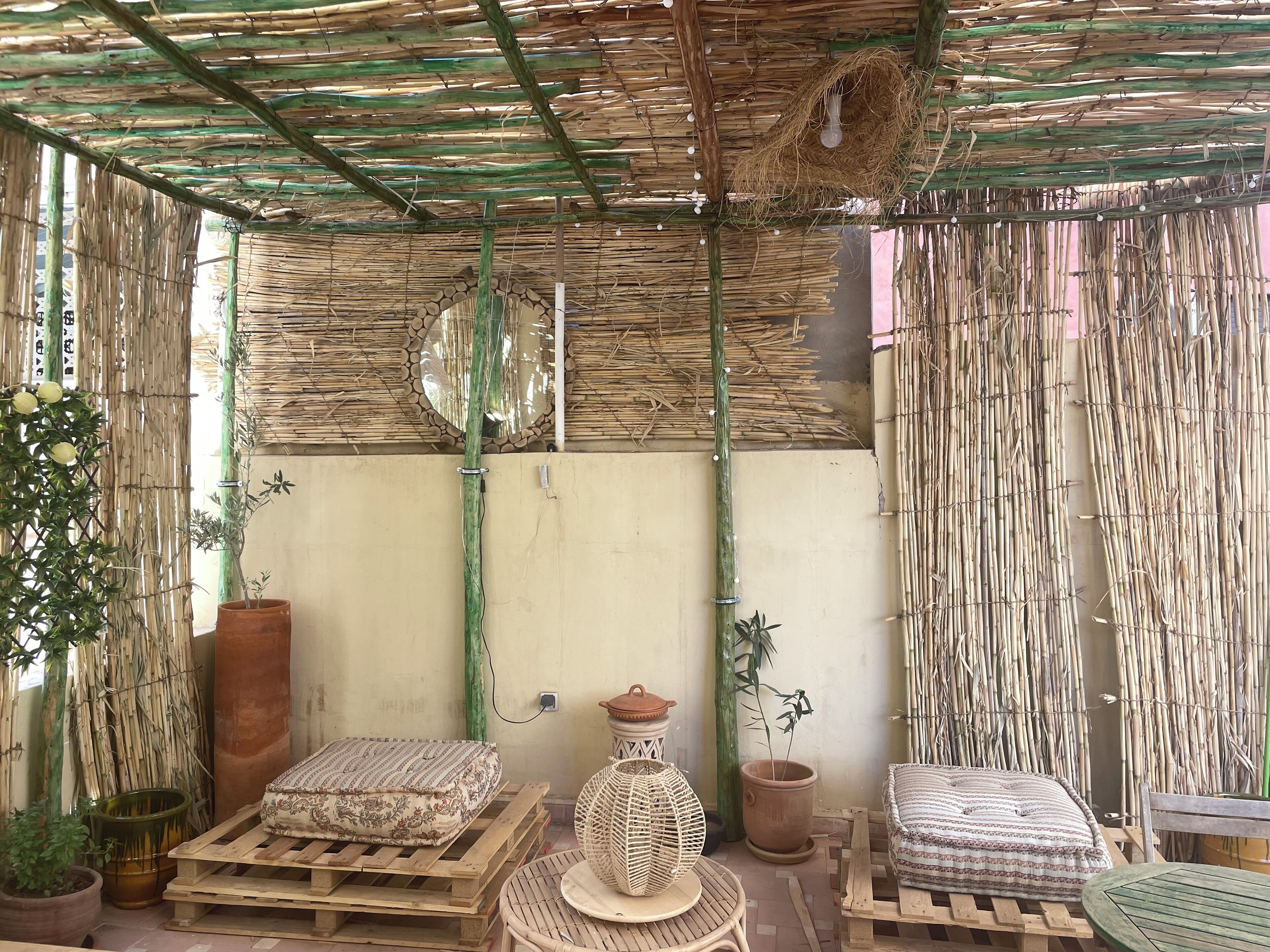 A room with a thatched roof, bamboo walls, and a hanging light fixture. There are two wooden beds with pillows, a woven chair, and a potted plant.