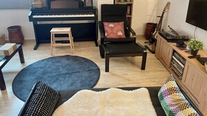 A living room with a piano, a black chair with a red pillow, a small wooden stool, a black rug, a white shag rug, and a wooden cabinet with a TV and a guitar. - Aux Nuits de Miel - Marrakech  (Marrakech)