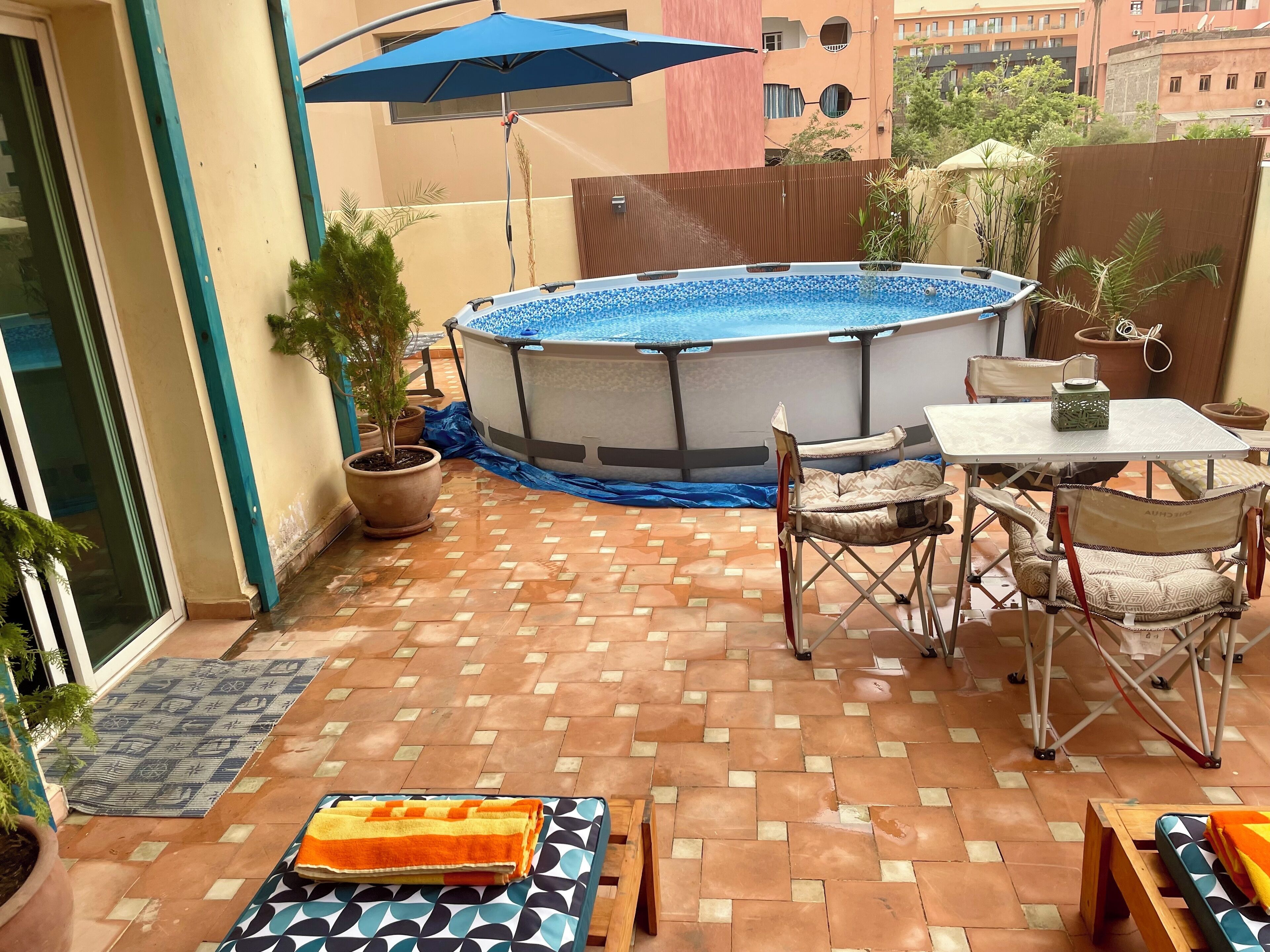 An outdoor patio with a swimming pool, a table with chairs, and potted plants.