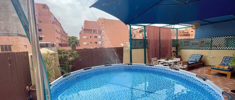 An outdoor swimming pool with a blue mosaic tile floor, surrounded by a wooden deck and a covered seating area with lounge chairs.