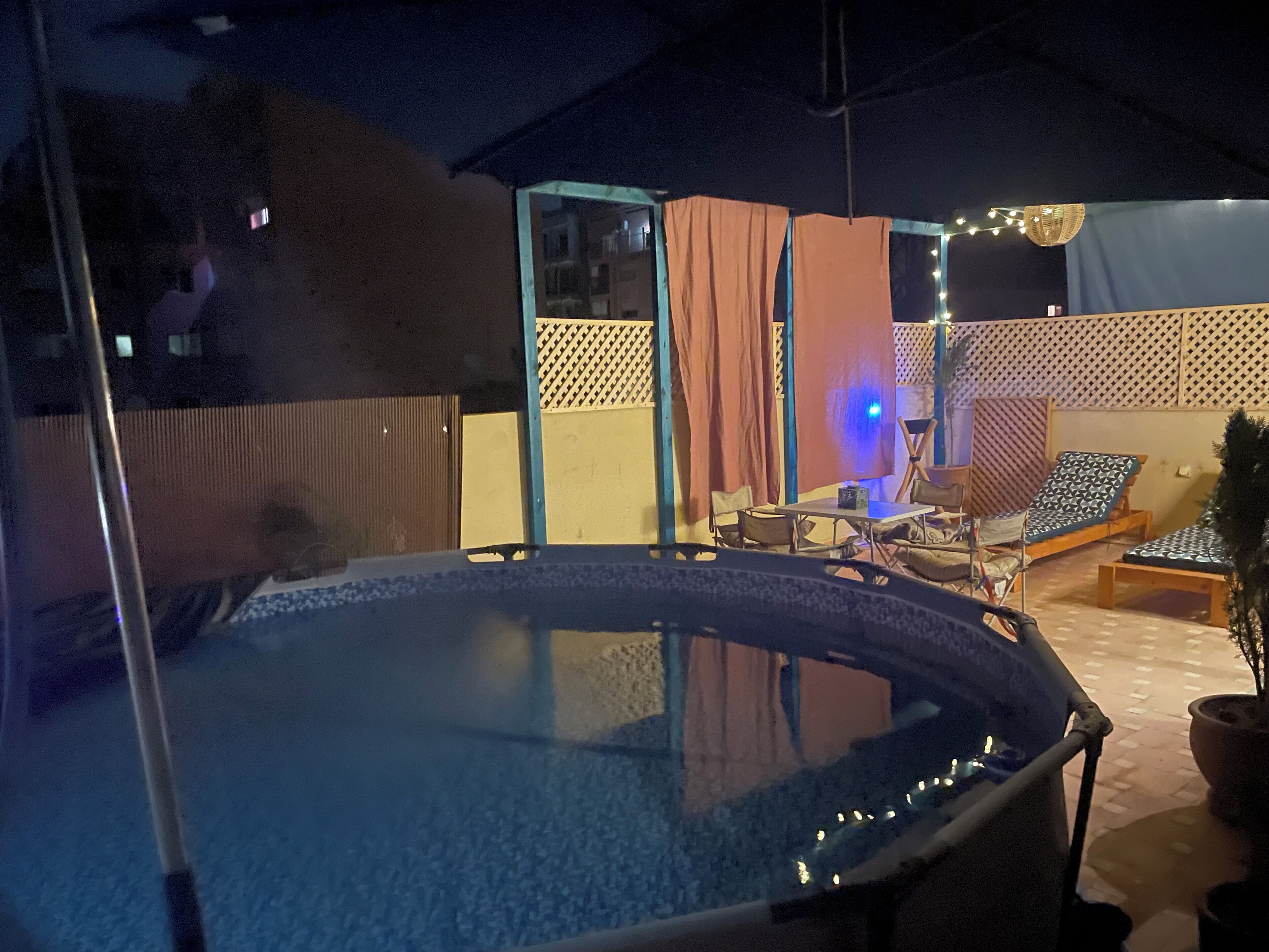 A hot tub area with a table and chairs, surrounded by a covered patio with curtains and string lights.