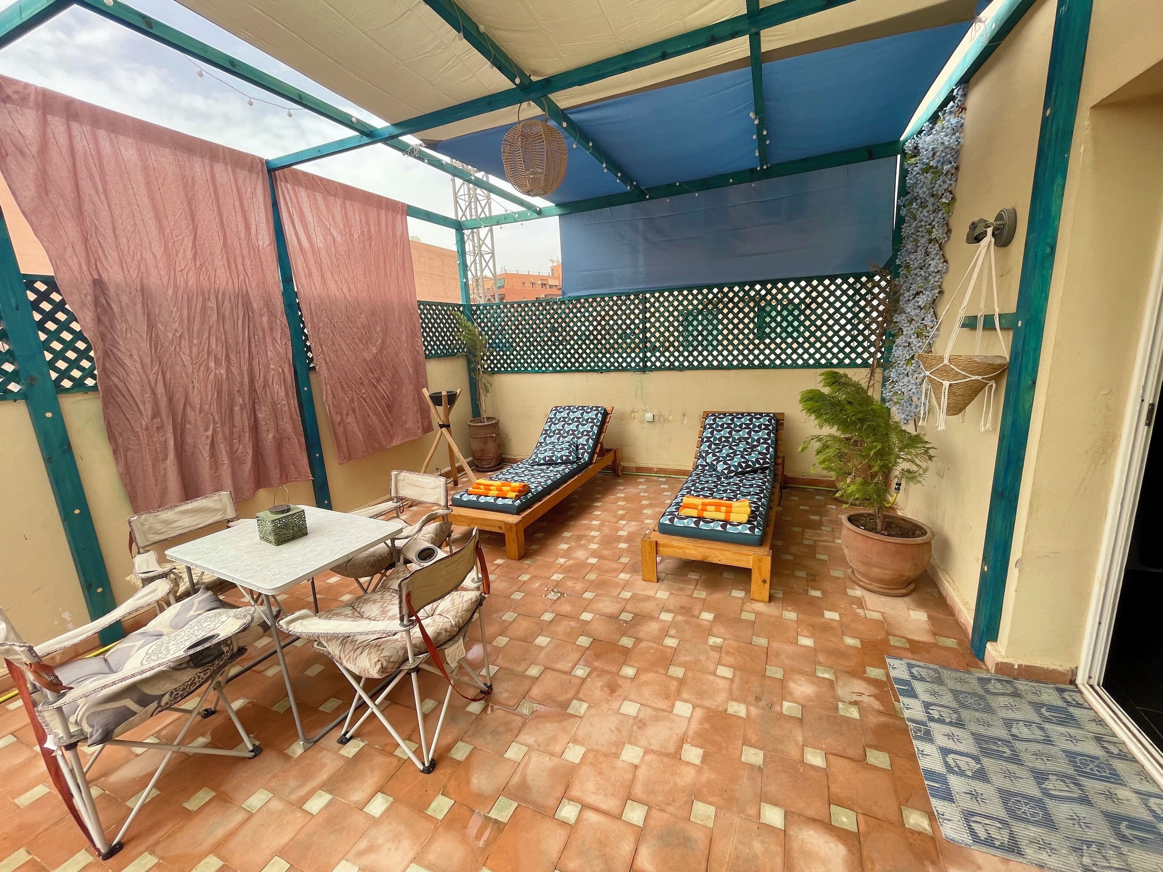 A sunlit patio with a table and chairs, two lounge chairs, a potted plant, and a decorative wall.