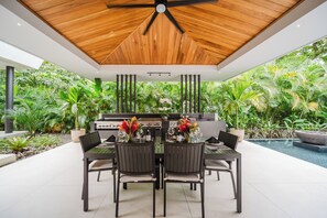 Un espace de restauration en plein air doté d’un plafond en bois, entouré d’une végétation luxuriante et d’une piscine.