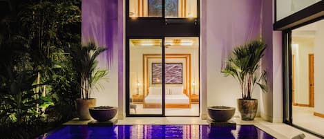 A hotel lobby with a pool, potted plants, and a view into a bedroom.