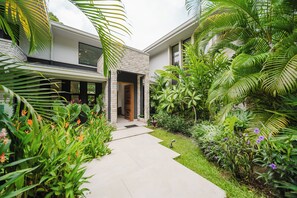 Une maison moderne avec une allée pavée menant à une porte en bois, entourée d’une végétation luxuriante et de plantes tropicales.