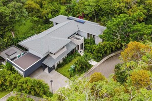 A modern two-story house with a flat roof, surrounded by lush greenery and a paved driveway.
