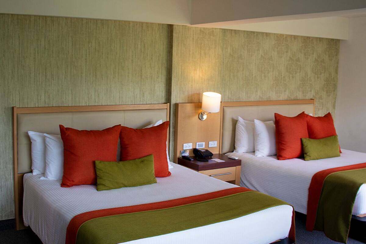 Two double beds with red and green pillows, a bedside table with a lamp, and a wall-mounted phone.