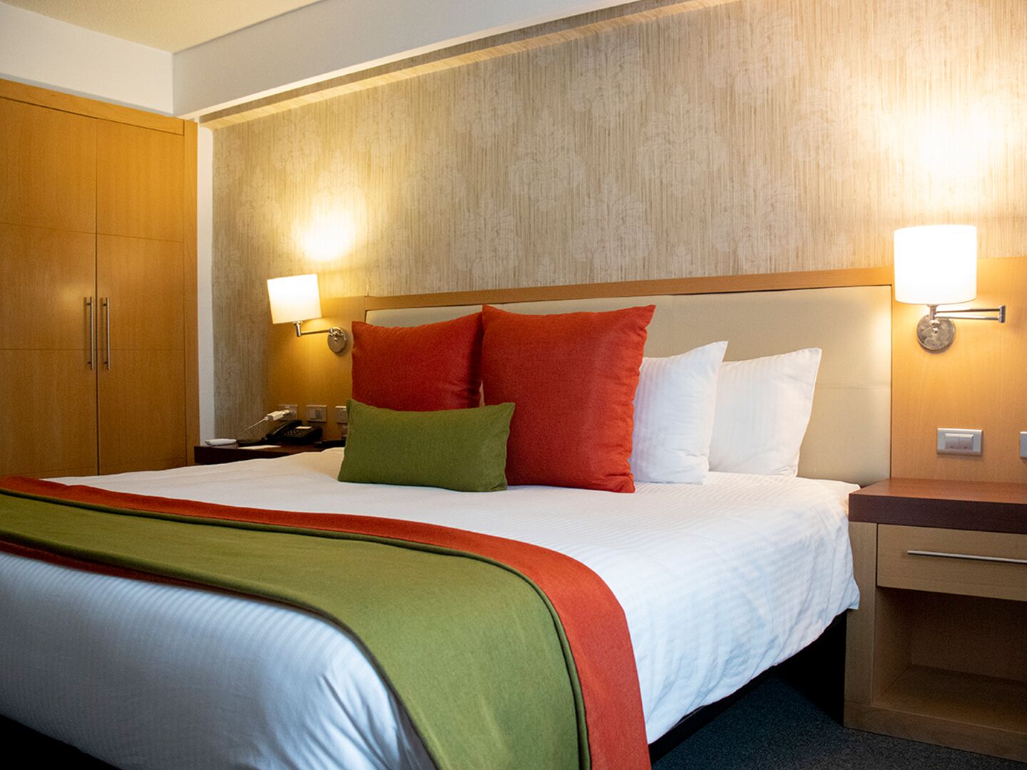 A hotel room with a large bed, two bedside tables, a wooden wardrobe, and wall-mounted lamps.