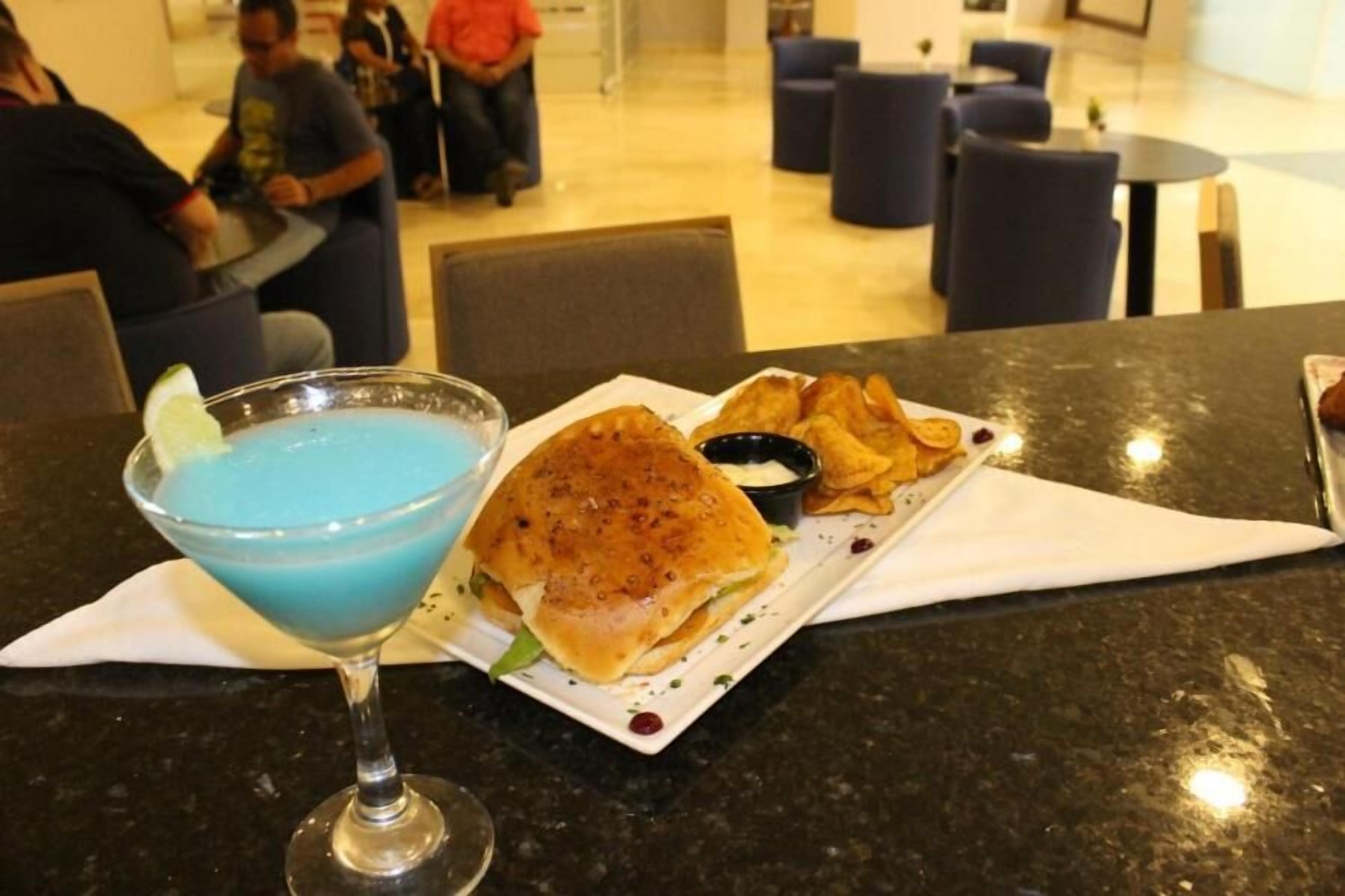 Et bord med en blå drink i et cocktailglas, en sandwich og en tallerken med pommes frites.