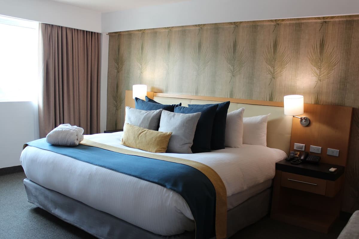 A hotel room with a large bed, a wooden headboard, two bedside tables with lamps, and a window with curtains.