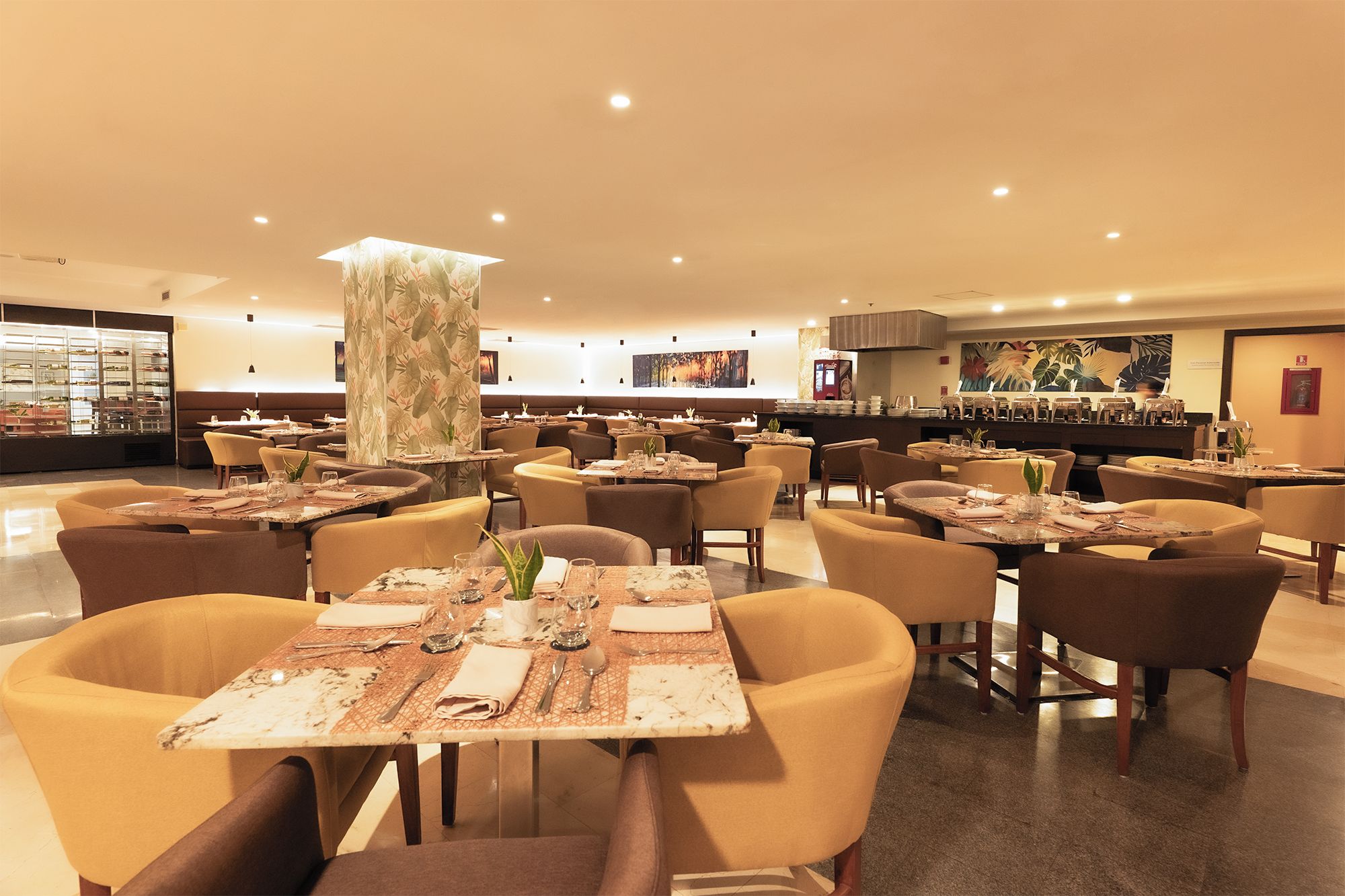 Un restaurante amplio con varias mesas y sillas, un pilar central decorado con motivos florales y una vitrina de postres al fondo.