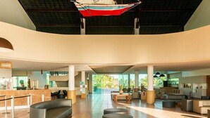 A spacious lobby with a check-in counter, seating areas, and a large window offering a view of greenery. - Apartamento Luxo no Beach Class Resort Muro Alto 3204 (Ipojuca)