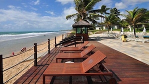 A wooden deck with lounge chairs and a thatched-roof structure overlooking a beach with palm trees and a clear sky. - Apartamento Luxo no Beach Class Resort Muro Alto 3204 (Ipojuca)