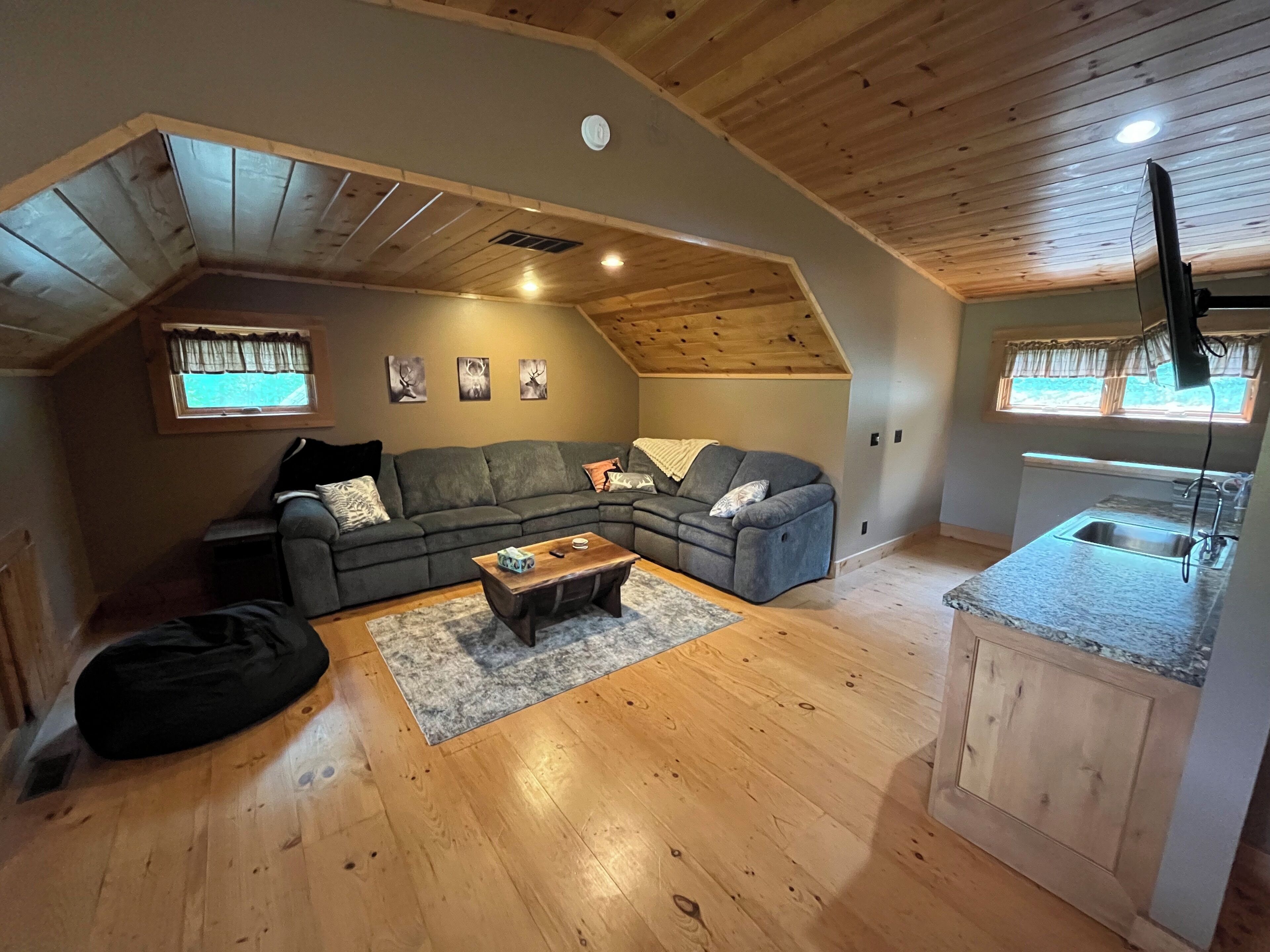 Un espacio habitable tipo loft con techo de madera, un sofá seccional gris, una mesa de centro y una cocina con encimera de granito.