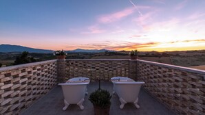 Un balcone in pietra con due vasche bianche, piante in vaso e vista su un tramonto su un paesaggio collinare.