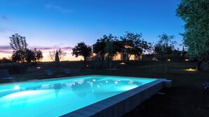 Una piscina rettangolare con superficie d'acqua cristallina e blu, circondata da un'area verde con alberi e una casa sullo sfondo.