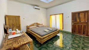 A room with a bed, a table with a bottle and tissues, a window with curtains, and a door. - Duyung Rooms and Resto by Zuzu (Kuta)