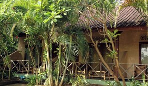 A building surrounded by lush tropical vegetation, with a tiled roof and a wooden fence. - Duyung Rooms and Resto by Zuzu (Kuta)