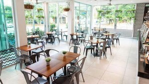 A spacious cafe with multiple tables and chairs, large windows offering a view of the outdoors, and hanging plants. - Soho Playa (Playa del Carmen)