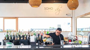 A bar area with a bartender preparing a drink, a variety of liquor bottles, and a sign that reads "SOHO PLATA". - Soho Playa (Playa del Carmen)